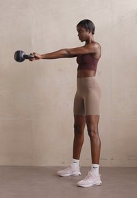 Egy nő bordó sportmelltartót és bézs rövidnadrágot visel, kezében egy fekete kettlebellt tart. Világos rózsaszín sportcipőt és fehér zoknit visel, semleges környezetben.