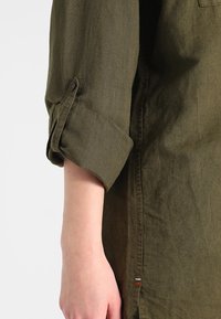 Camicia di lino verde oliva con maniche arrotolate, dotata di polsino con bottone e di una fessura laterale con un accento a striscia colorata. Tessuto testurizzato.
