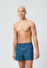 Jonge man met kort blond haar, gekleed in blauwe en groene geruite boxershorts, staande tegen een effen lichtgrijze achtergrond.
