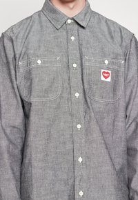 Grijs overhemd met knopen, twee borstzakken, witte knopen en een rode hartvormige logo patch met de tekst "Carhartt WIP" op de rechterborstzak.