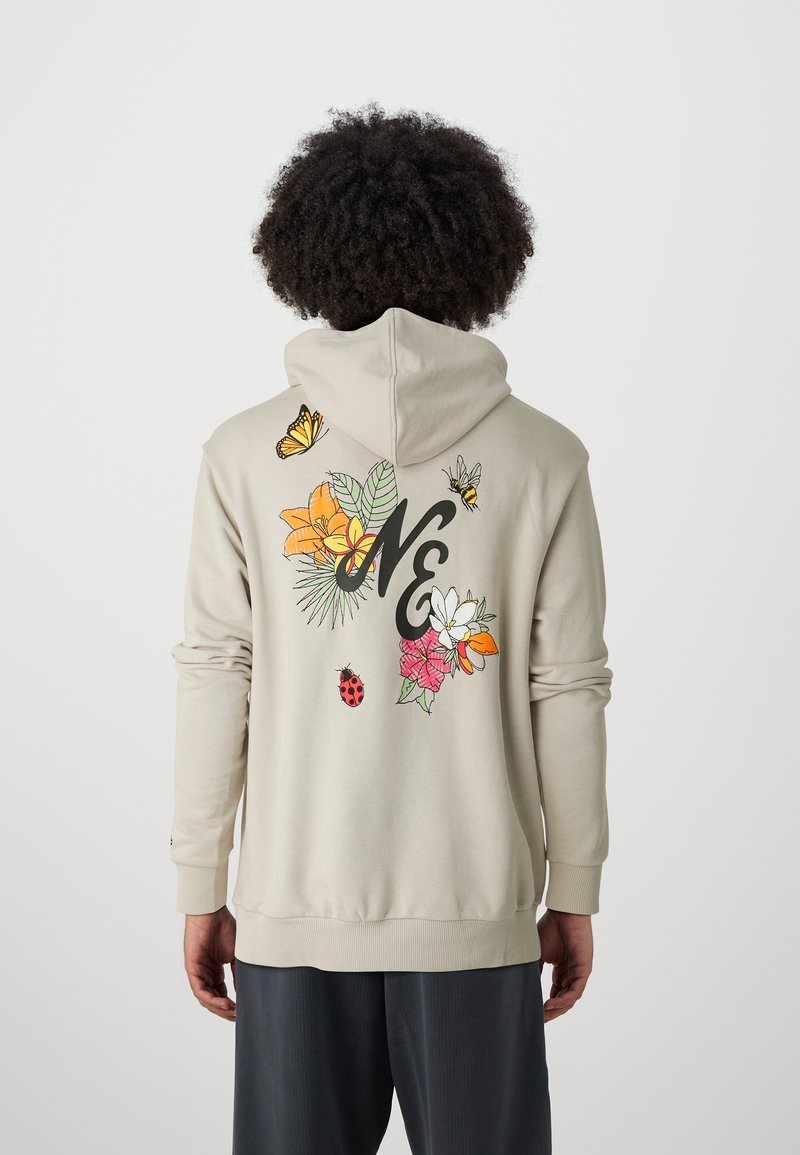 Persona con cabello rizado que lleva una sudadera beige con diseños de flores, mariposas, abejas y mariquitas, y letras negras "NE" en la parte posterior.