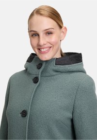 Grøn, tekstureret frakke med hætte i sort. Har store knapper og en høj krave. Stoffet ser varmt og blødt ud, velegnet til køligere vejr.