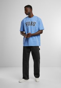 T-shirt din bumbac de culoare albastru deschis, cu un logo negru, îndrăzneț "FUBU", asociat cu pantaloni cargo negri și adidași albi,Stand pe un fundal neutru.