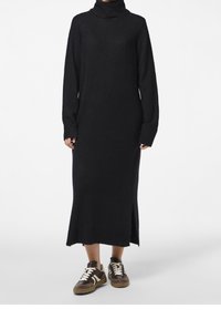 Abito a collo alto nero realizzato in un materiale morbido, con maniche lunghe, vestibilità comoda e uno spacco laterale all'orlo. Abbinato a sneaker marroni.