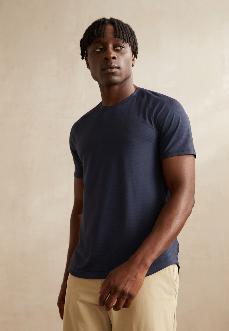 Chemise navy à manches courtes avec un col rond, réalisée en tissu lisse, présentant un motif texturé et de légers fentes sur les côtés au niveau de l'ourlet.