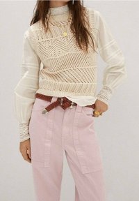Suéter de punto crema con patrón geométrico, superpuesto a una blusa blanca con mangas fruncidas; combinado con pantalones de pierna ancha rosa claro y cinturón trenzado.
