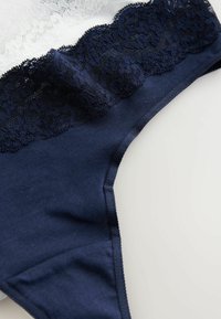 Tamnoplava tanga gaćice s čipkastim pojasom sa valovitim rubom. Mekana tkanina s glatkom teksturom i minimalnim detaljima šivanja.