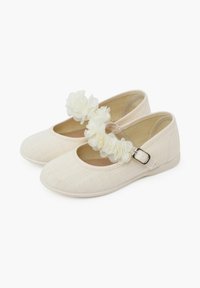 Zapatos de tela beige con punta redondeada, que presentan una correa en el tobillo y adornos de flores blancas. Suela plana y textura suave.