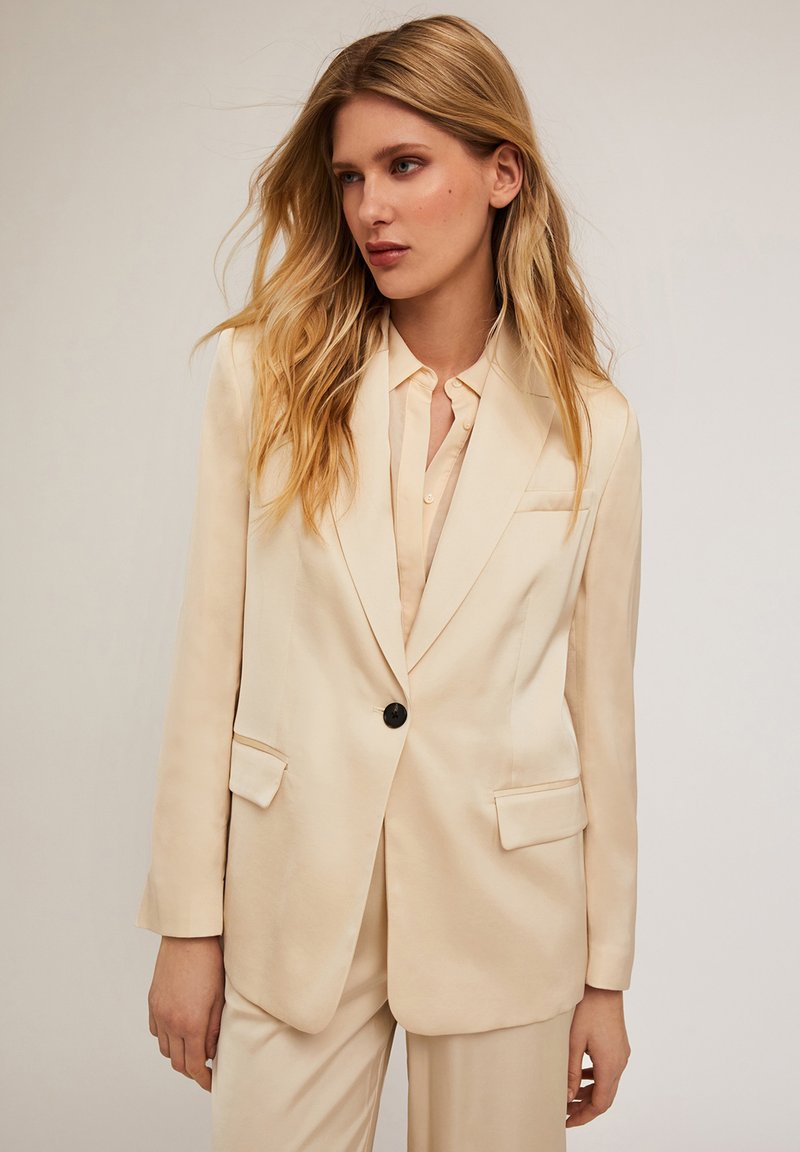 Sacou bej cu o textură netedă, asemănătoare satinului, având un singur nasture negru, rever tăiat și două buzunare frontale.