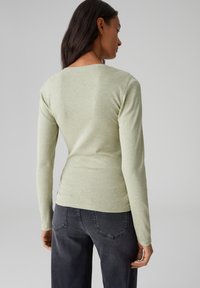 Hellgrünes Langarm-T-Shirt aus weichem Stoff, mit Rundhalsausschnitt und taillierter Passform, kombiniert mit dunkelgrauen Jeans.