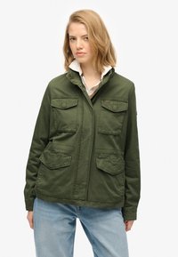 Groene katoenen jas met een rits- en knoopsluiting, twee borstzakken, twee lagere zakken en een staande kraag. Draag een witte blouse eronder.