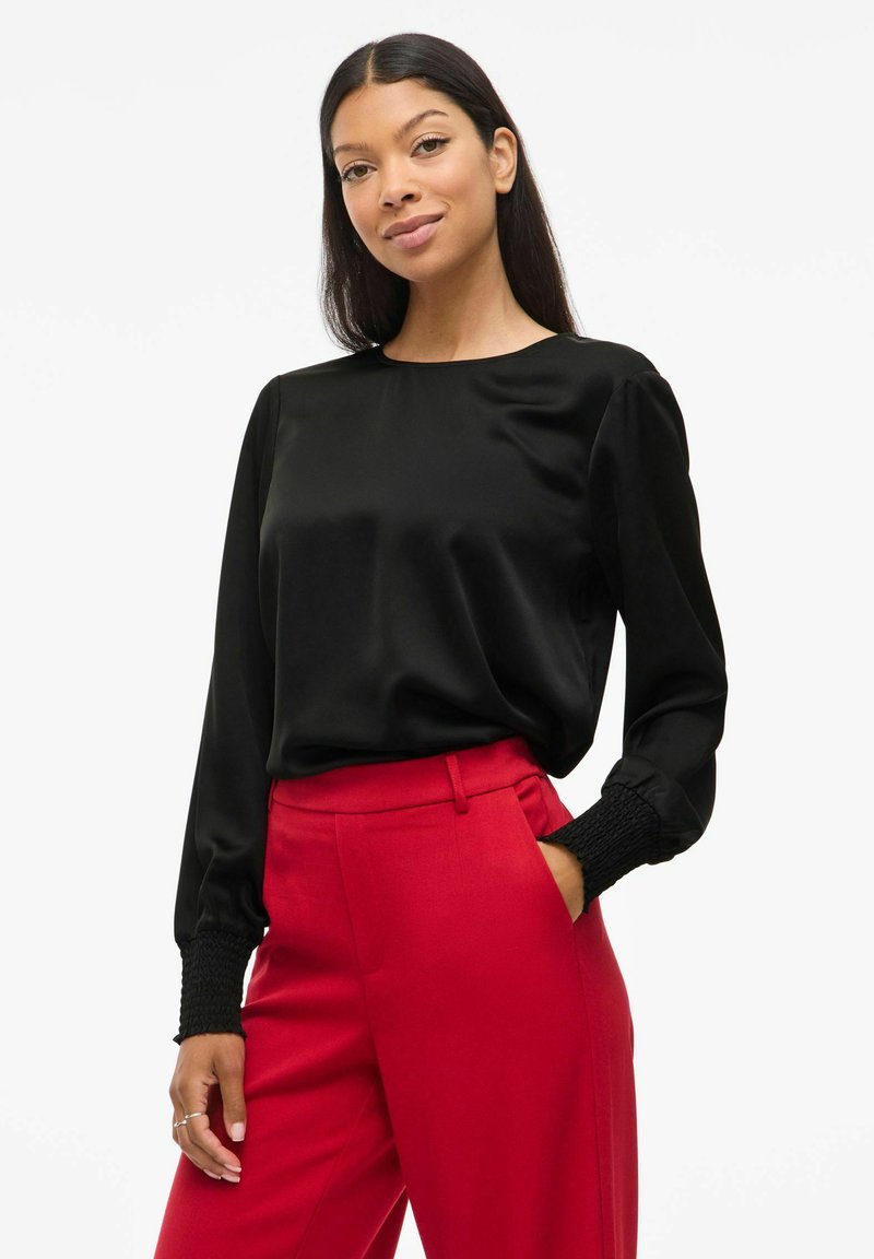 Blusa nera in satin con maniche lunghe e polsini arricciati, abbinata a pantaloni rossi a vita alta. Texture liscia, classico scollo rotondo.