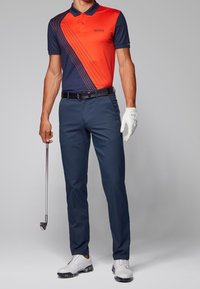 Muži v námořnickém a červeném diagonálním pruhovaném polo tričku, s námořnickými kalhotami, bílou golfovou rukavicí, držící golfovou hůl a v bílých golfových botách, na šedém pozadí.