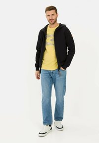 Sort zip-up hoodie med ribbet tekstur, båret over en gul grafisk t-shirt, parret med lyseblå jeans og hvide sneakers med sorte accenter.