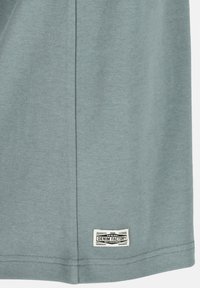 Ljus teal bomullstyg med en slät textur. Har en synlig varumärkesetikett sydd vid fållen. Enkelt mönster med rak skärning.