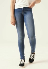 Jeans de denim azul ajustados con una textura suave, cintura media y un ligero desvanecimiento. Combinados con zapatillas negras que tienen cordones blancos.