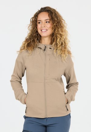 Lächelnde Frau mit lockigem Haar, die eine beige Jacke mit Reißverschluss und Reißverschlusstaschen sowie blaue Hosen trägt, steht mit den Händen in den Taschen.