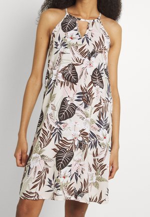 Vrouw draagt een beige mouwloze jurk met bruine, zwarte, groene en roze tropische blad- en bloemmotieven en een klein voorste sleutelgat.