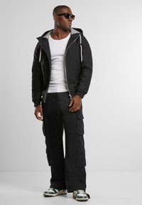 Svart zip-up jakke med grå innside og snorhette, kombinert med svarte cargo-bukser med flere lommer og sneakers med grønne detaljer.