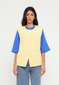 Veste sans manches en polaire jaune avec deux poches avant, associée à un pull bleu à manches courtes. Pantalon en denim coupe droite.