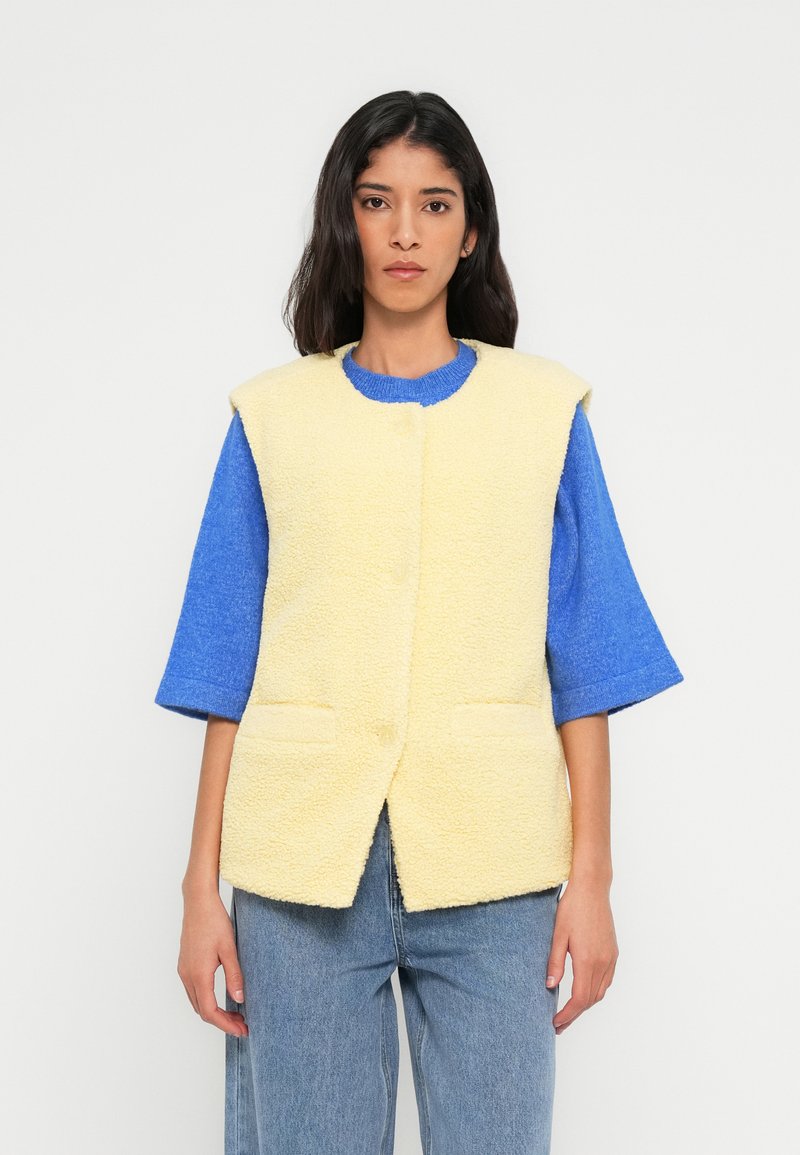 Veste sans manches en polaire jaune avec deux poches avant, associée à un pull bleu à manches courtes. Pantalon en denim coupe droite.