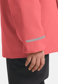Korallenfarbene wasserdichte Jacke mit glatter Textur, ausgestattet mit einem grauen reflektierenden Streifen am Bund. Schwarze Hose teilweise sichtbar.