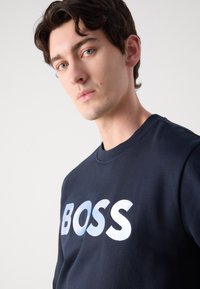 Jūras spēku pelēks džemperis, kas izgatavots no kokvilnas, ar izteiktu gradienta "BOSS" logo. Dizains ietver noapaļotu kakla izgriezumu un garām piedurknēm.