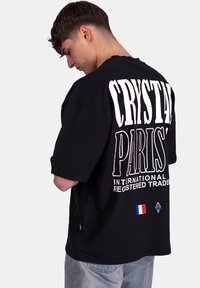 T-shirt noir en coton avec un grand texte blanc « CRYSTAL PARIS ». Comprend un petit drapeau français et un logo de diamant sur le côté.
