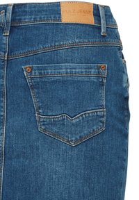 Detalje af baglomme på blå denimjeans med brun syning og en tan læder-mærkat med mærket "Pulz Jeans" på linningen.