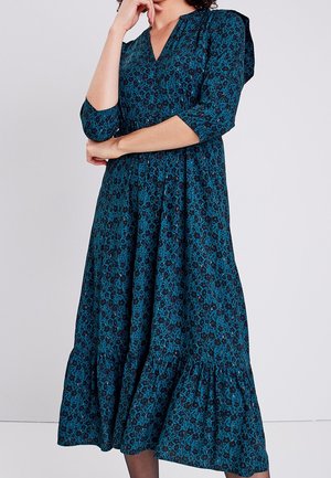 Donna con un vestito midi verde acqua con motivo floreale nero, maniche a tre quarti e orlo arricciato, posa con una mano sul mento.