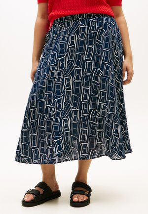 Γυναίκα με midi φούστα μπλε με σχέδια, κόκκινο τοπ και μαύρα πέδιλα ανοιχτά στη μύτη με ρυθμιζόμενα λουράκια, να στέκεται σε λευκό φόντο.