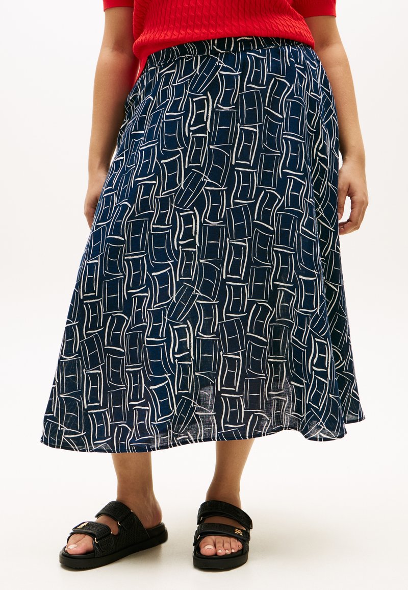 Γυναίκα με midi φούστα μπλε με σχέδια, κόκκινο τοπ και μαύρα πέδιλα ανοιχτά στη μύτη με ρυθμιζόμενα λουράκια, να στέκεται σε λευκό φόντο.