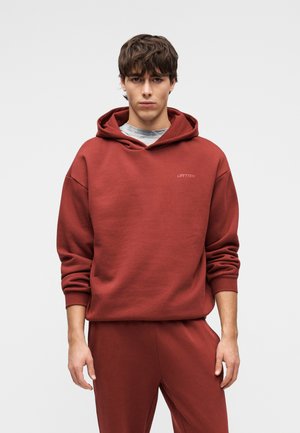 Ung mand med brunt hår iført en rustfarvet hættetrøje og matchende bukser, stående mod en ensfarvet lys baggrund.