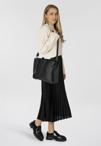 Bolso de mano de cuero negro con un diseño estructurado, dobles asas superiores y una correa para el hombro removible. Lleva puesto un suéter ligero y una falda negra plisada.