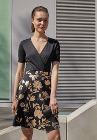 Haut noir à manches longues associé à une jupe évasée, ornée de motifs floraux dorés. L'ensemble est complété par un petit sac à main noir.