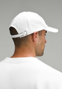 Vit baseballkeps med en böjd skärm och silverfärgad justerbar rem bak, bärs av en person i en vit skjorta.