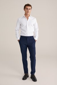 Witte blouse, marineblauwe dresspants en zwarte schoenen, staande tegen een neutrale achtergrond. Het shirt heeft een klassieke kraag en een getailleerd ontwerp.