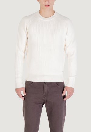 Maglione a coste color crema con collo rotondo e maniche raglan. Il materiale sembra a maglia; abbinato a pantaloni grigio scuro. Nessun motivo visibile.