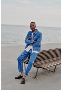 Denim jakna svijetlog pranja, bijela košulja ispod, plave traperice i tamnosmeđe cipele. Model sjedi na drvenoj klupi pored vode.
