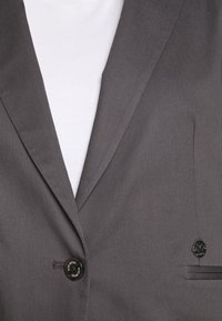 Gros plan sur un blazer gris ajusté avec un seul bouton et une petite épingle logo en métal sur une chemise blanche côtelée.