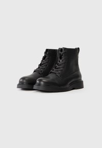 Botas de tobillo de cuero negro con un acabado texturizado, punteras redondas, parte delantera con cordones y una suela de goma resistente. Presenta una cremallera corta en el lateral.