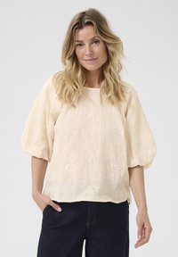 Blouse beige clair brodée avec des manches bouffantes et un col rond. Motifs floraux et finition texturée. Associée à un pantalon bleu marine.