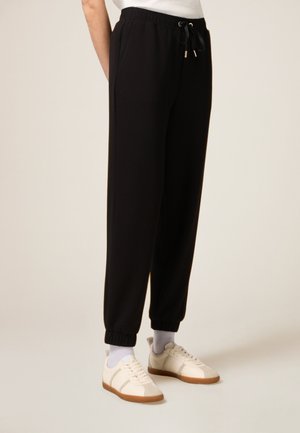 Black jogger pants with elastic waistband and drawstring. Features tapered ankles and side pockets. Worn with light-colored sneakers.