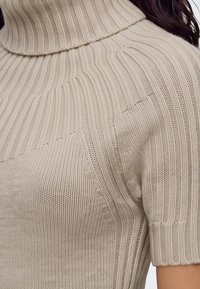 Gros plan d’un pull col roulé en maille côtelée beige à manches courtes porté par une personne aux cheveux foncés.