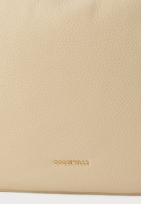 Τσάντα χειρός από δέρμα μπεζ με υφή. Διαθέτει χρυσό ανάγλυφο λογότυπο "Coccinelle" στην κάτω γωνία. Απλός και μοντέρνος σχεδιασμός.