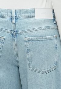 Ανοιχτό μπλε τζιν παντελόνι με πίσω τσέπες και λευκή ετικέτα μάρκας. Το ύφασμα έχει κλασική υφή τζιν και ελαφριά ξεθώριασμα.