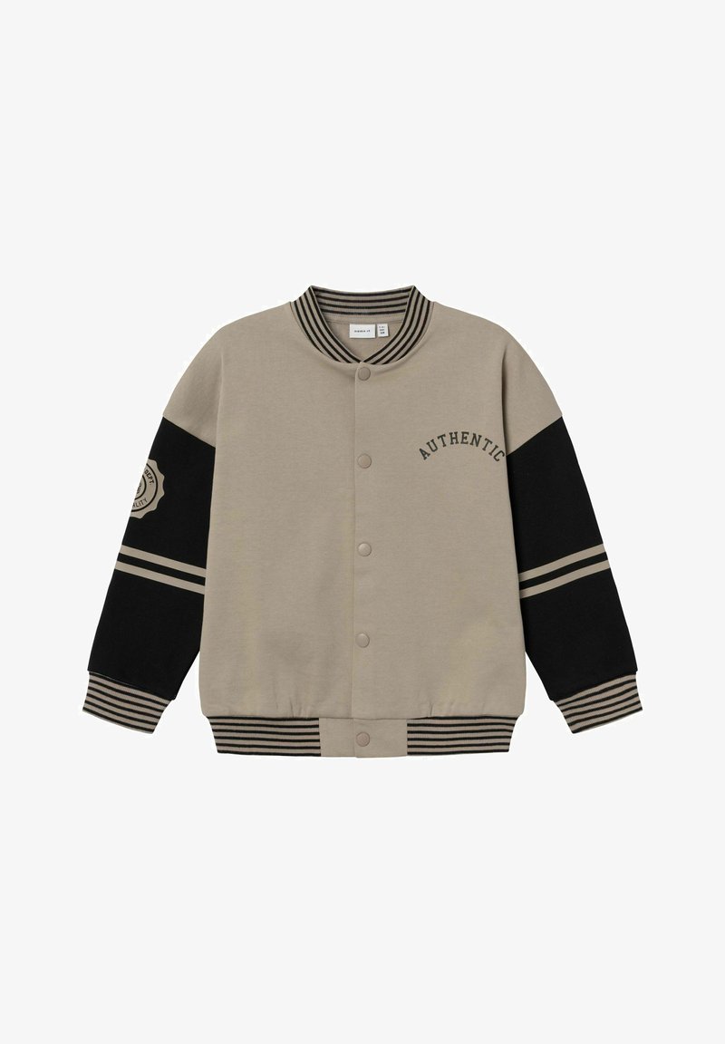 Veste universitaire beige avec manches noires. Présente un col et des poignets côtelés, des boutons pressions, et le texte "AUTHENTIC" au-dessus du côté gauche.