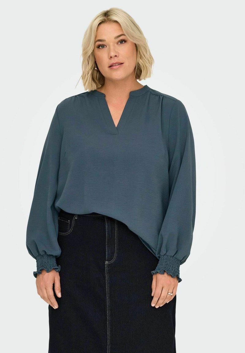 Blouse teal avec un décolleté en V et de longues manches bouffantes avec des poignets froncés. Associée à une jupe en denim affichant une couture à l'avant.