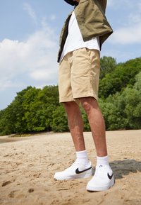 Shorts beiges à coupe décontractée, associés à des baskets blanches ornées d'un swoosh noir, et des chaussettes blanches jusqu'à la cheville, sur une plage de sable.