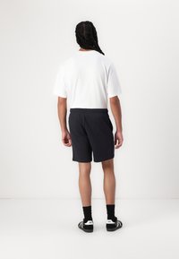 White t-shirt, black shorts, elastic waistband, and black sneakers with white accents. View from the back, standing on a plain background.